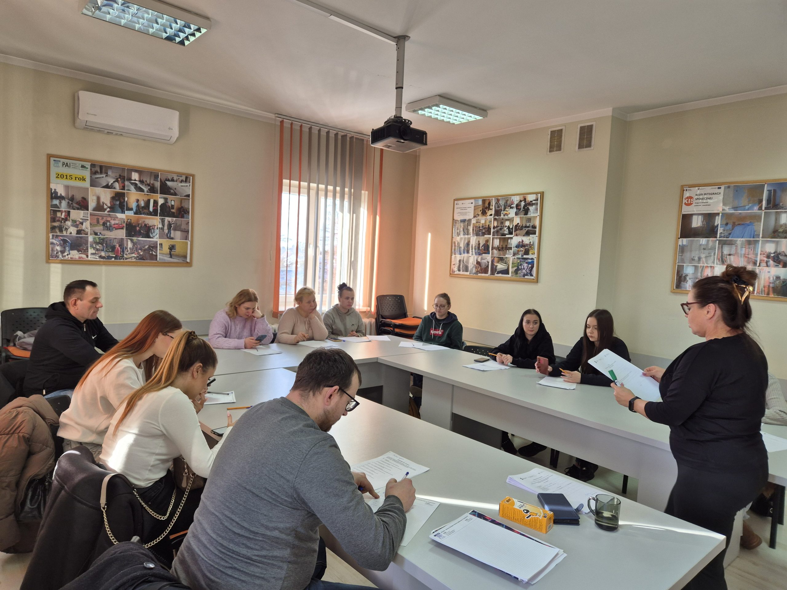 Na zdjęciu widzimy uczestników szkolenia pracujących nad dokumentami oraz instruktorkę czytającą treść dokumentu, która stoi na środku sali
