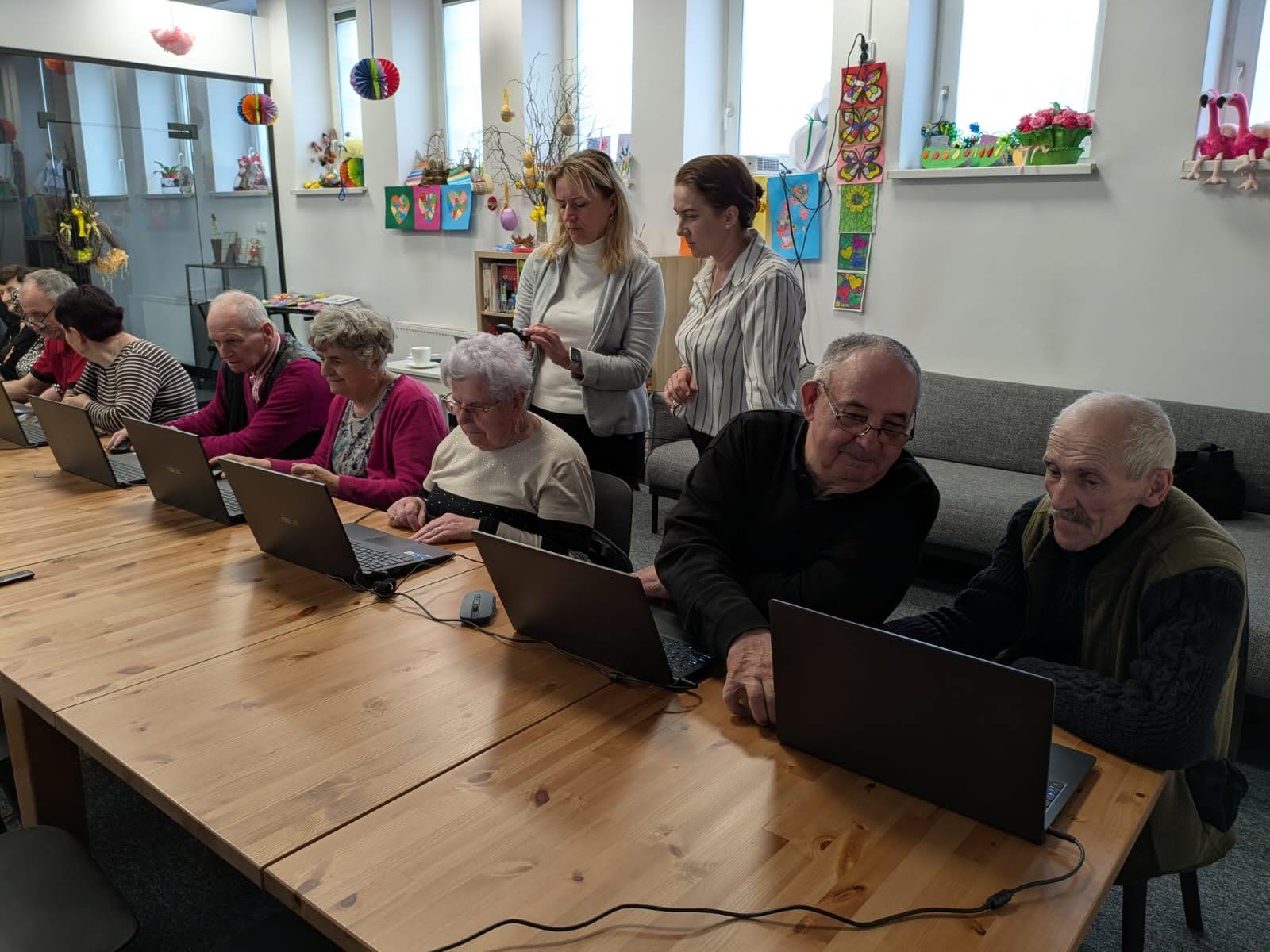 3) Na fotografii instruktorka, terapeutka oraz seniorzy siedzący przy komputerach.