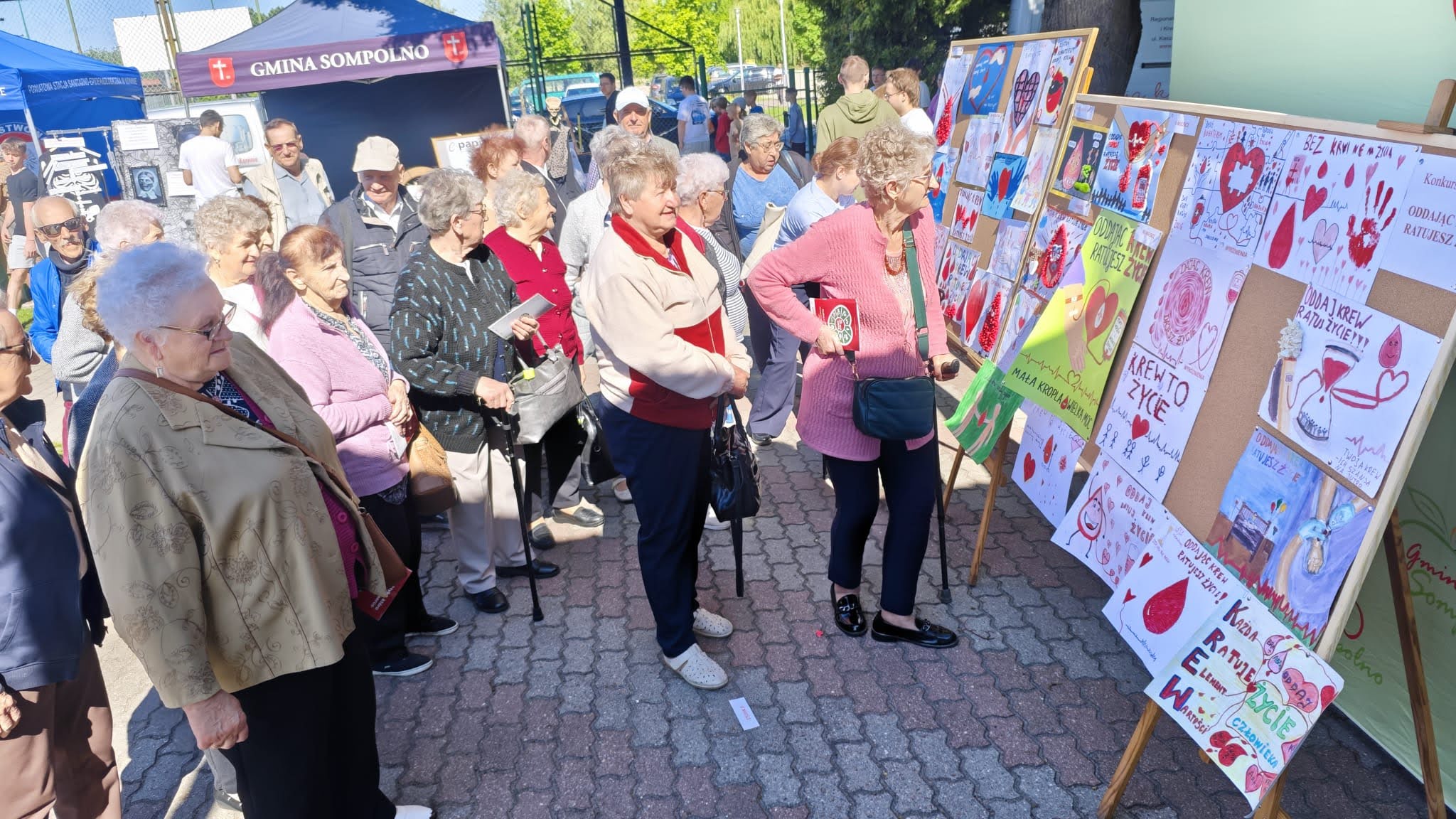 1) Zdjęcie wykonane przy szkole podstawowej w Sompolnie. Przedstawia uczestników domu dziennego pobytu oglądających wystawę prac plastycznych na temat krwiodawstwa.