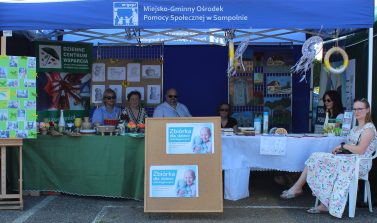 1) Zdjęcie wykonane podczas Pikniku Sołtysów w Sompolnie. Przedstawia stoisko Domu Dziennego Pobytu oraz Dziennego Centrum Wsparcia w Sompolnie. Pod namiotem dyrektor i pracownicy MGOPS oraz nauczycielka ze szkoły podstawowej w Sompolnie.