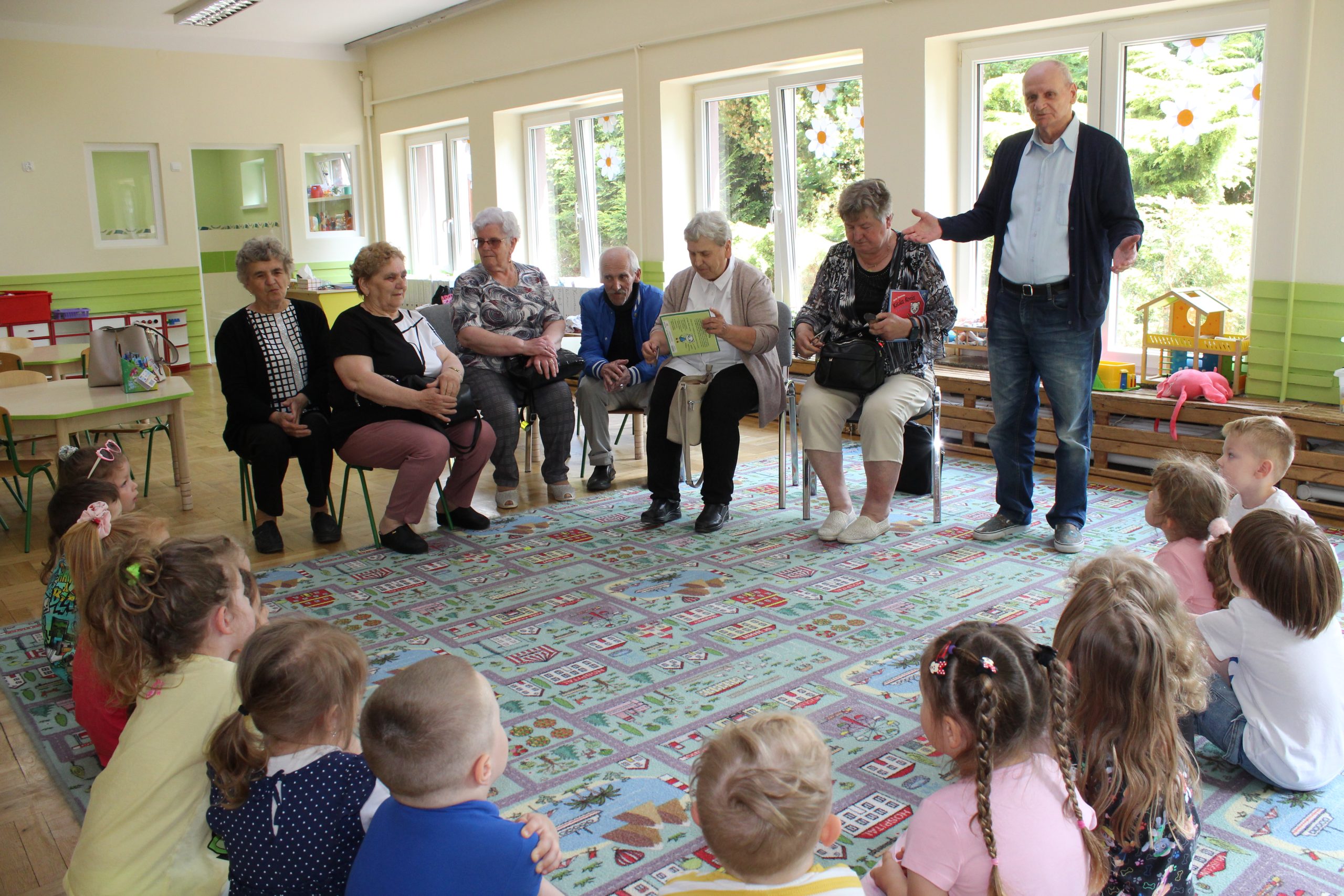 1) Zdjęcie wykonane w przedszkolu miejskim Zielony Zakątek w Sompolnie, w grupie świerszczy. Przedstawia seniorów siedzących na krzesłach i przedszkolaków siedzących na dywanie.