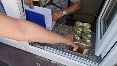 1) Zdjęcie wykonane przed budynkiem MGOPS w Sompolnie. Przedstawia okno przez które wydawana jest żywność. Widać ręce chwytające miód w słoikach oraz ręce osoby przygotowującej kartę do podpisu.