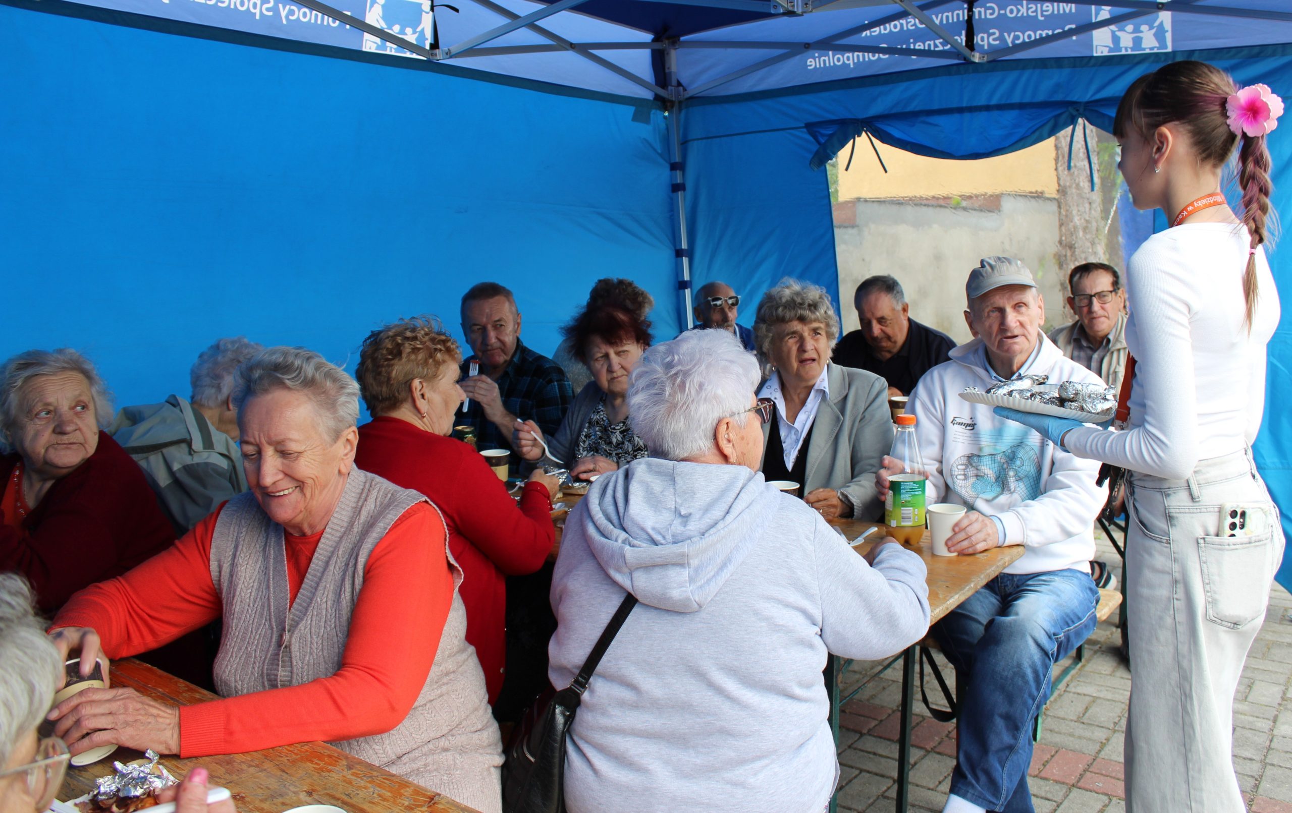 Na zdjęciu seniorzy z Domu Dziennego Pobytu siedzący pod niebieskim namiotem podczas spotkania przy grillu.