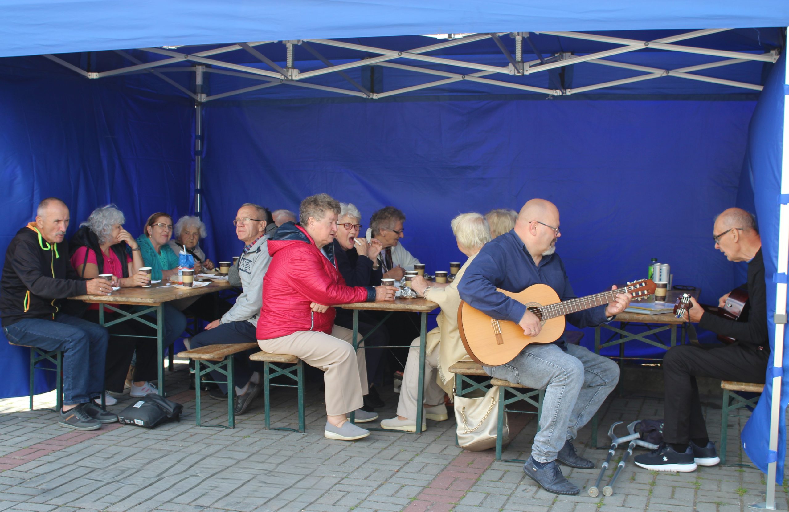 Na zdjęciu seniorzy z Dziennego Centrum Wsparcia siedzący pod niebieskim namiotem podczas spotkania przy grillu. Po prawej stronie dwaj mężczyźni siedzący przy stole i grający na gitarach.