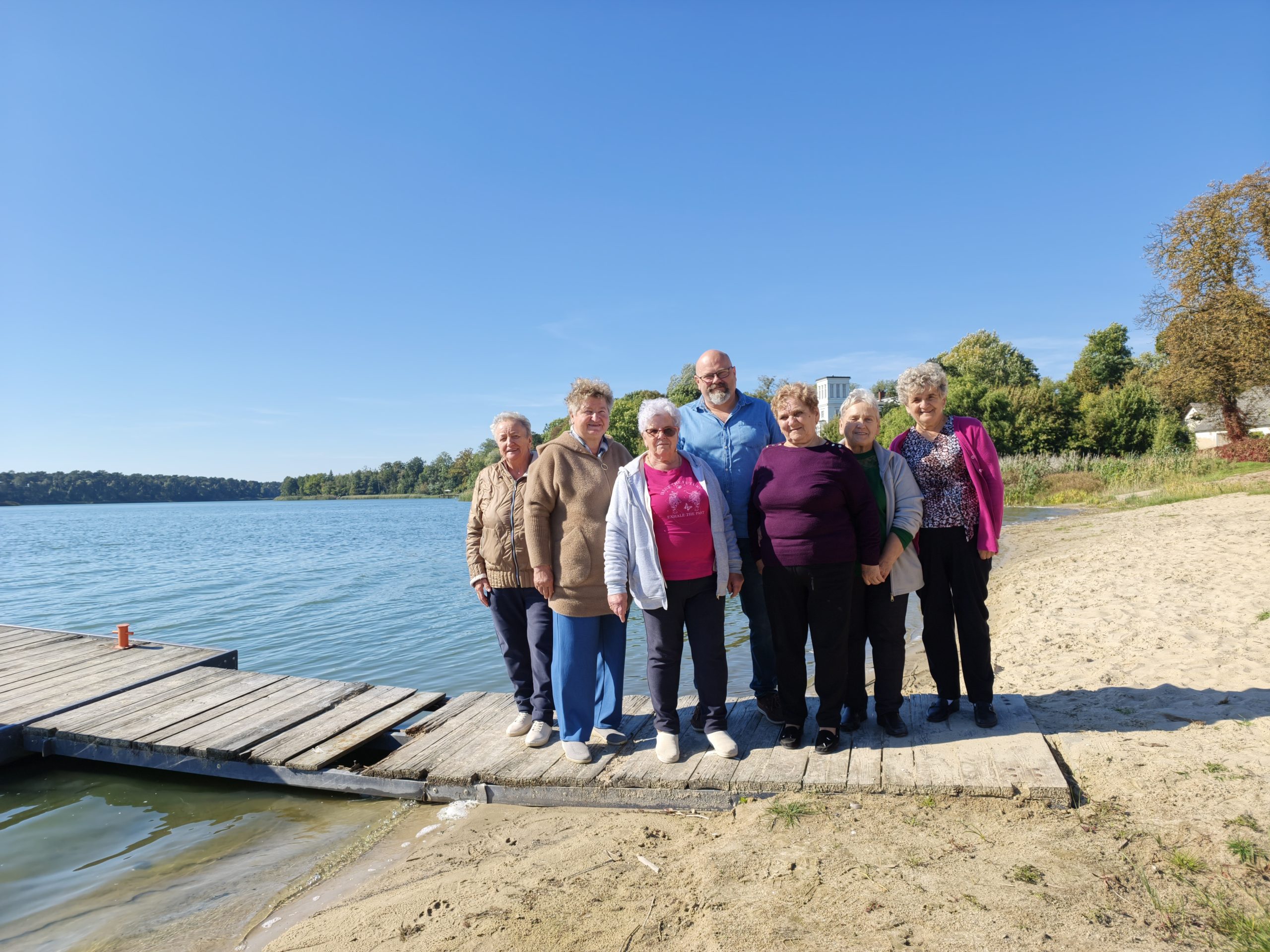 1. Zdjęcie wykonane nad jeziorem w Lubstowie. Przedstawia grupę seniorów wraz z Dyrektorem Miejsko-Gminnego Ośrodka Pomocy Społecznej