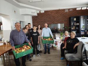 6. Zdjęcie wykonane w sali głównej DDP. Przedstawia delegację Koła Wolontariatu z Mąkoln wraz z opiekunami oraz seniorów z DDP, którzy odbierają otrzymane od dzieci jabłka.