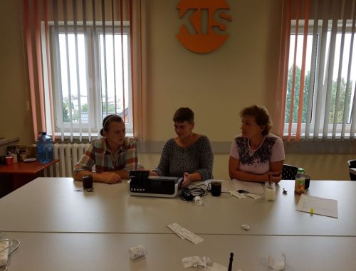 Na zdjęciu znajdują się trzy osoby siedzące przy stole. Przed nimi stoi kasa fiskalna i leżą paragony.