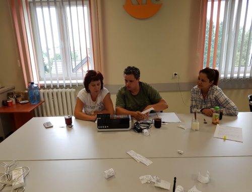 Na zdjęciu znajdują się trzy osoby siedzące przy stole. Przed nimi stoi kasa fiskalna i leżą paragony.