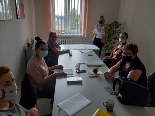 Na zdjęciu znajduje się 6 kobiet. Pięć z nich siedzi przy stole, na którym leżą dokumenty, telefony komórkowe i staja szklanki. Jedna Kobieta stoi z dokumentami w ręku. Wszyscy mają na twarzach maseczki.