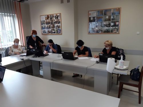 Na zdjęciu znajduje się pięć kobiet siedzących przy stole. Przed każdą stoi otwarty laptop. Jedna kobieta pochyla się przy jednym z laptopów. Wszyscy maja na twarzach maseczki.