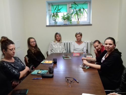 Na zdjęciu znajduje się sześć osób. Wszyscy siedzą przy stole, na którym leżą zeszyty, długopisy i okulary.