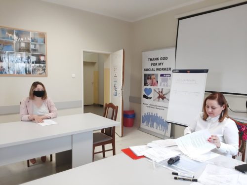 Na zdjęciu znajdują się dwie osoby siedzące przy dwóch stołach. Jedna z osób ma na twarzy maseczkę.