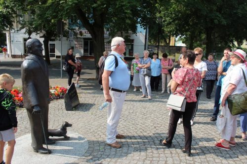 Na zdjęciu widać grupę osób w różnym wieku biorących udział w wycieczce do Ciechocinka. W centralnym miejscu mężczyzna – przewodnik, a z lewej strony pomnik Jerzego Waldorffa.