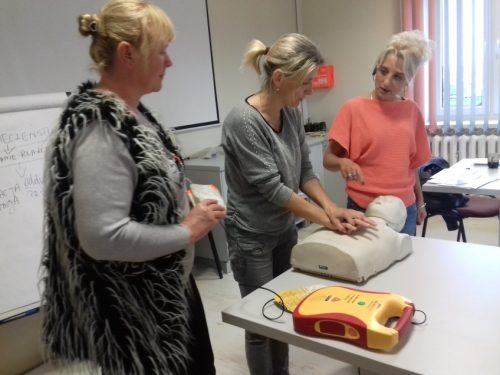 Na zdjęciu znajdują się trzy kobiety. Na stole leży tułów manekina. Jedna z kobiet uczy pozostałe techniki masażu serca.