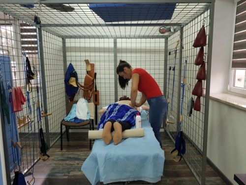 Na zdjęciu pomieszczenie, w którym znajduje się stół do masażu. Na stole leży kobieta, którą masuje rehabilitantka.