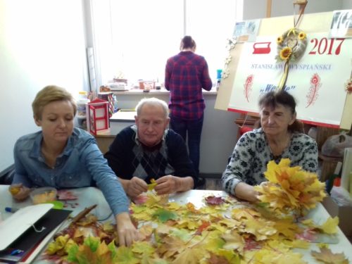 Na zdjęciu znajdują się cztery osoby. Trzy osoby siedzą przy stole, na którym leży dużo suszonych liści. Jedna osoba stoi przy oknie tyłem do pozostałych.