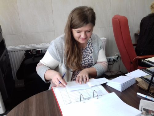 Na zdjęciu znajduje się jedna kobieta siedząca przy biurku. Przed nią leży segregator z dokumentami. Kobieta coś pisze.