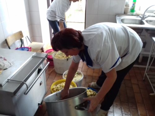 Na zdjęcia znajdują się dwie osoby w białych fartuchach. Jedna osoba pochylona myje duży garnek. na podłodze stoją pojemniki z resztkami jedzenia.